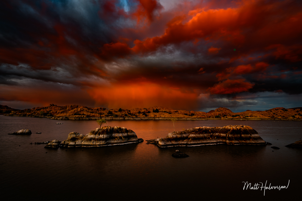 Fiery red and orange clouds dramatically fill the sky over a tranquil lake, with rugged rock formations scattered across the water's surface. The stark contrast between the darkening storm and the illuminated clouds creates a stunning visual impact. Shot slightly from the left. 