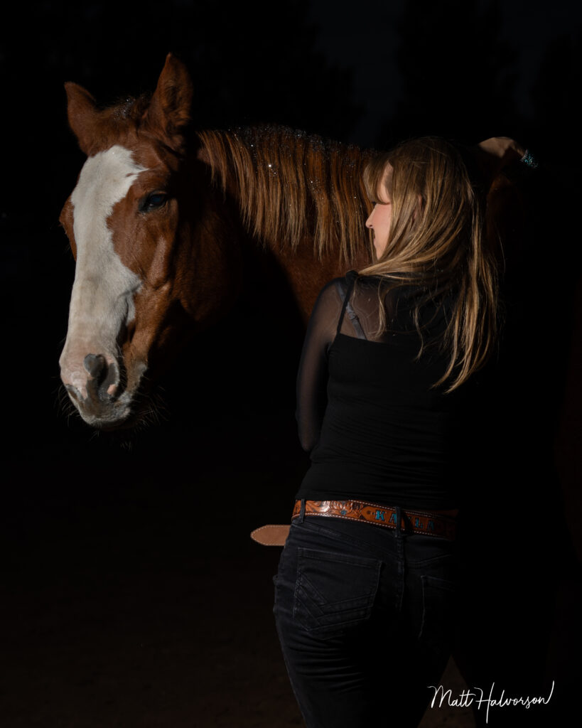 A woman with long blonde hair embraces a brown horse with a white blaze, both softly lit against a black background to highlight their emotional connection.