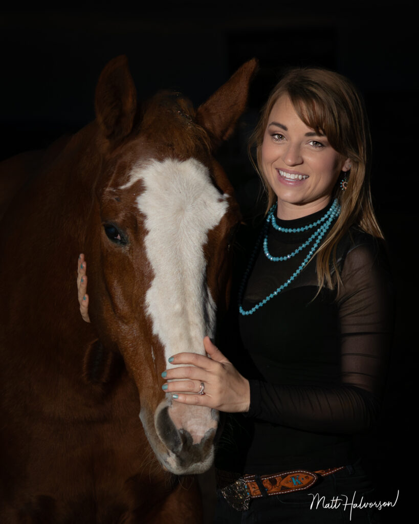 A smiling woman stands beside a brown horse with a white blaze, gently touching its face as both are illuminated against a dark background in a fine‑art portrait style
