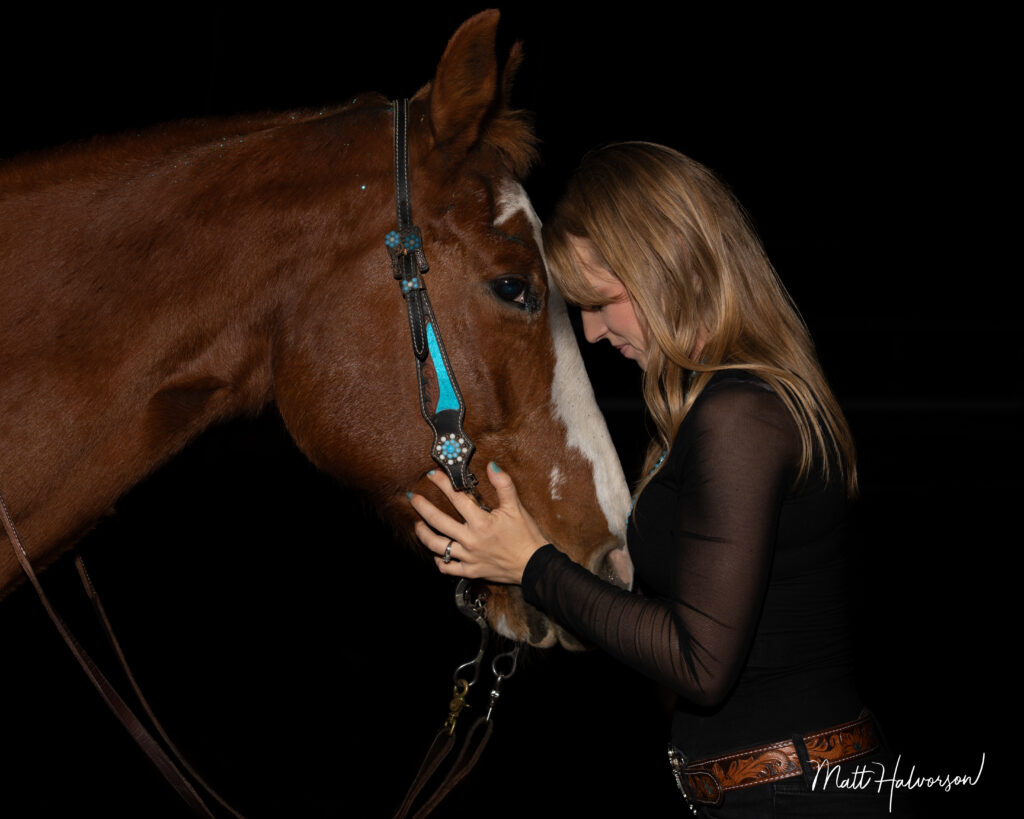 A woman stands with her head against a brown horse's head both framed against a dark background in a calm, fine‑art equine portrait.